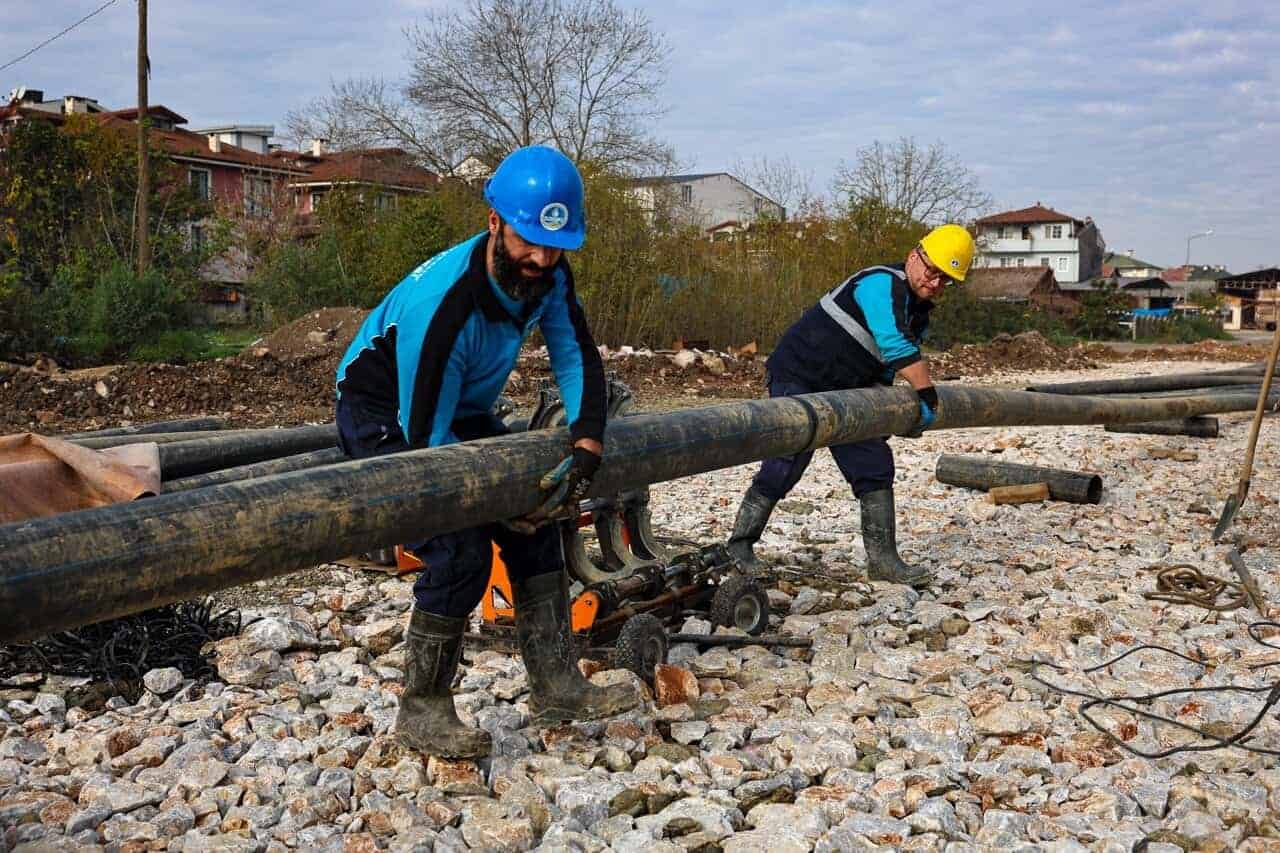 İki işçi, özel güvenlik önlemleriyle kanalizasyon borusu döşüyor, inşaat sahasında çalışma ortamı.