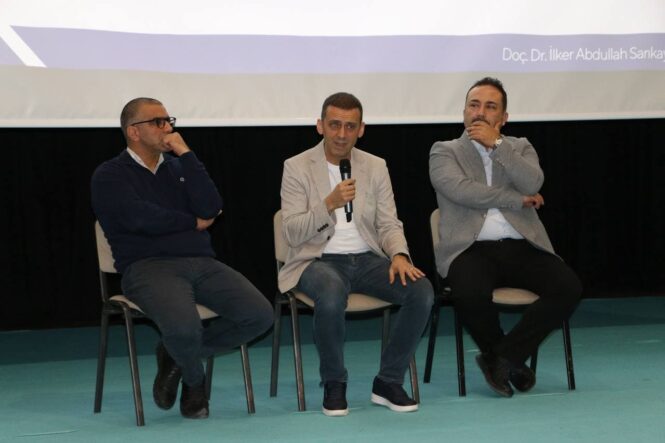 Basın merkezinde üç kişi panelde konuşma yapıyor, arka planda projeksiyon ekranı var.